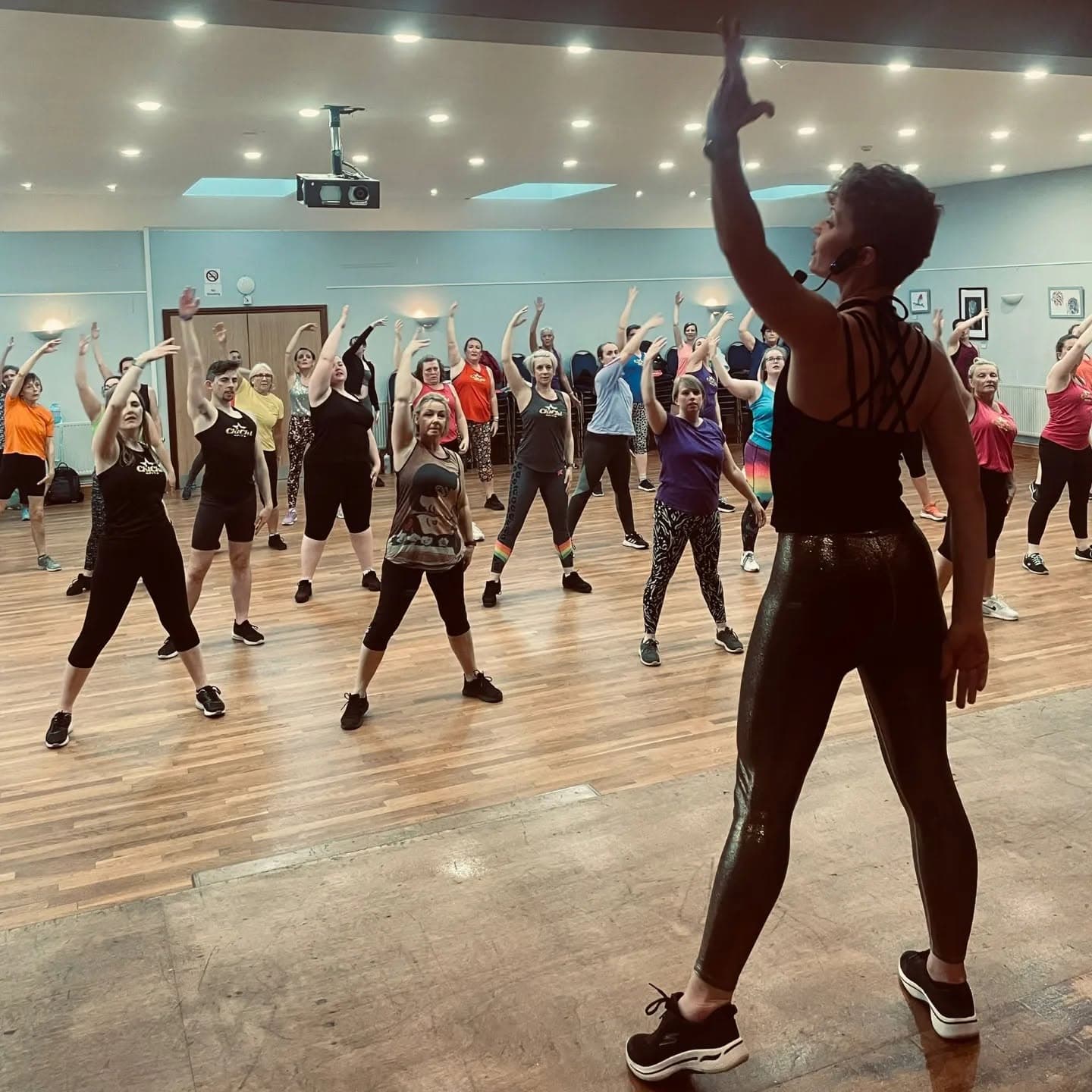 Instructeur leidt een volle ChiChi Fit zaal met opgeheven armen — cardio, choreografie en showenergie